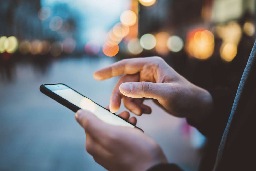 A male uses his right hand to scroll through a page on a smartphone. You can see city lights and streets blurred in the background.