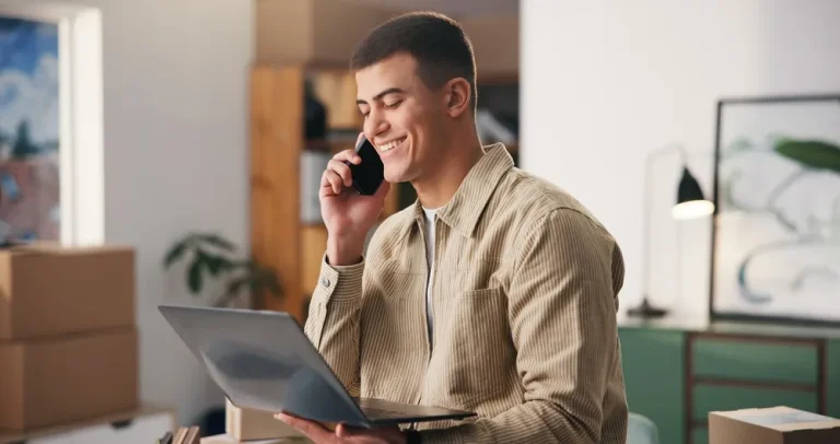 Young man smiling on the phone while looking at his laptop in his home