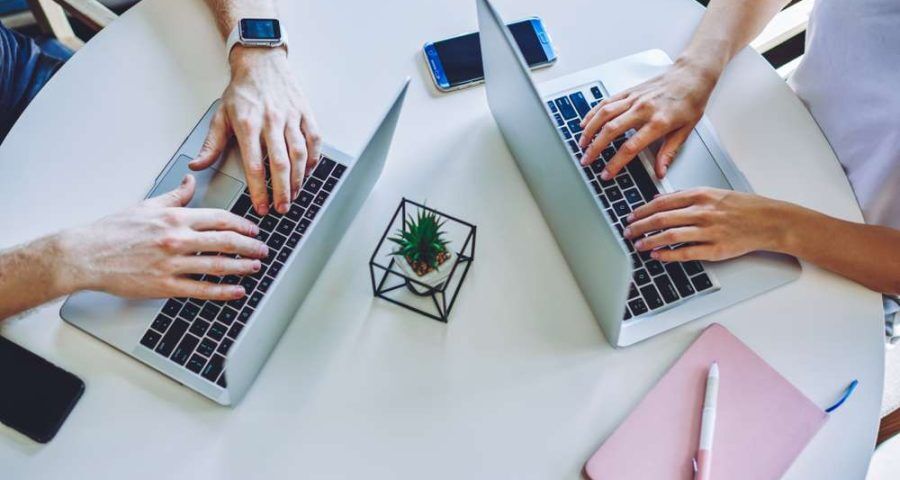 two people typing on their laptops on a white table with a flower in the middle, with a phone also on the table