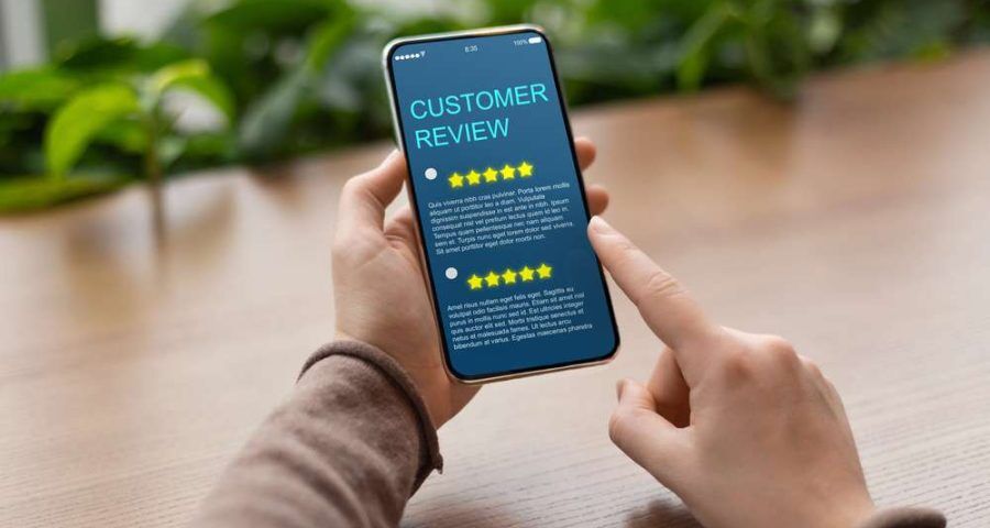 A person's hands are shown scrolling through a smartphone screen on a wooden table. The screen includes a customer reviews page with several five star reviews on it.