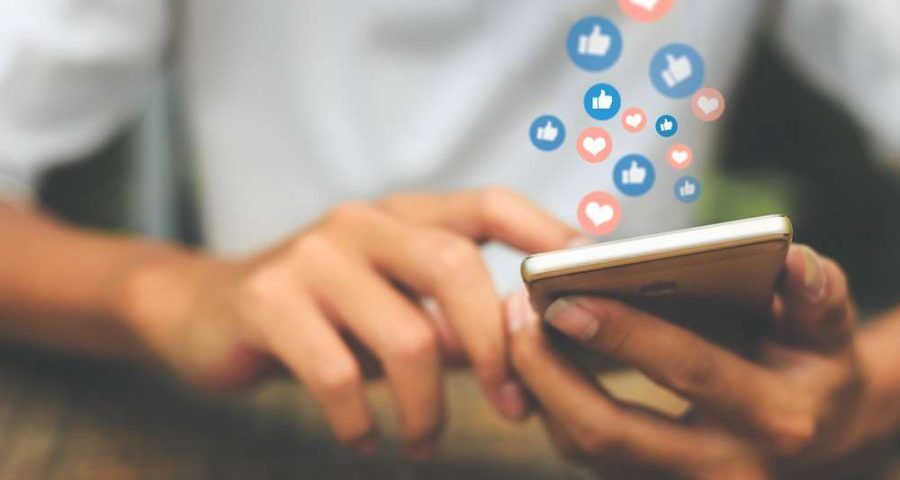 A mans hands can be seen operating a smart phone. Hearts and thumbs up symbols appear from the phone as a way to visualize social media engagement.