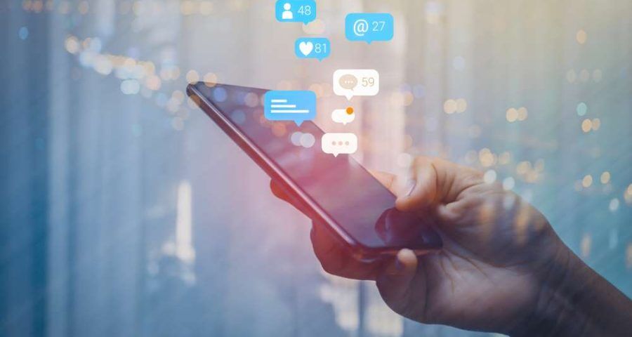 A close up of a person's hand holding a smartphone. Several thought bubbles with social media numbers can be seen above the phone screen. These are meant to symbolize social media impressions.