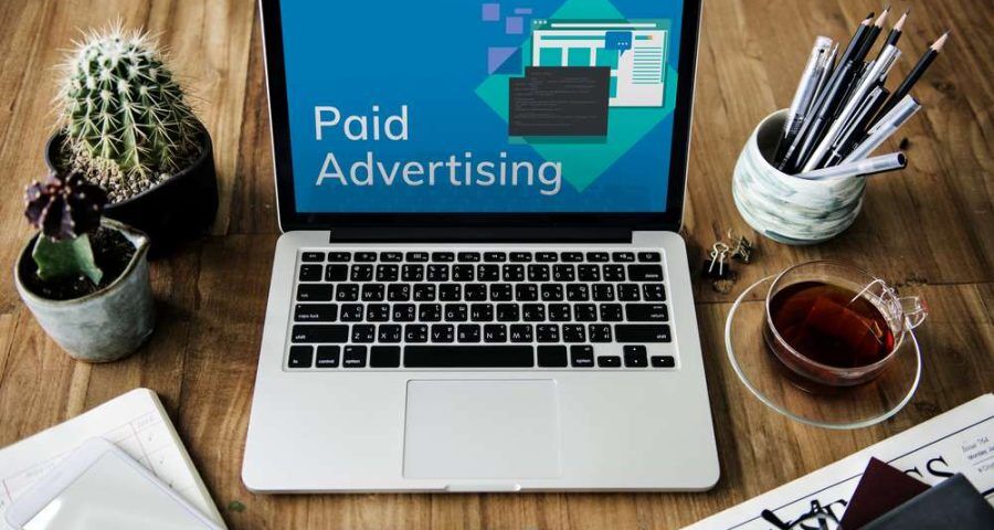 A laptop computer with the words "paid advertising" printed on the screen. You can see several sheets of paper, potted plants and a mug of coffee surrounding the laptop.